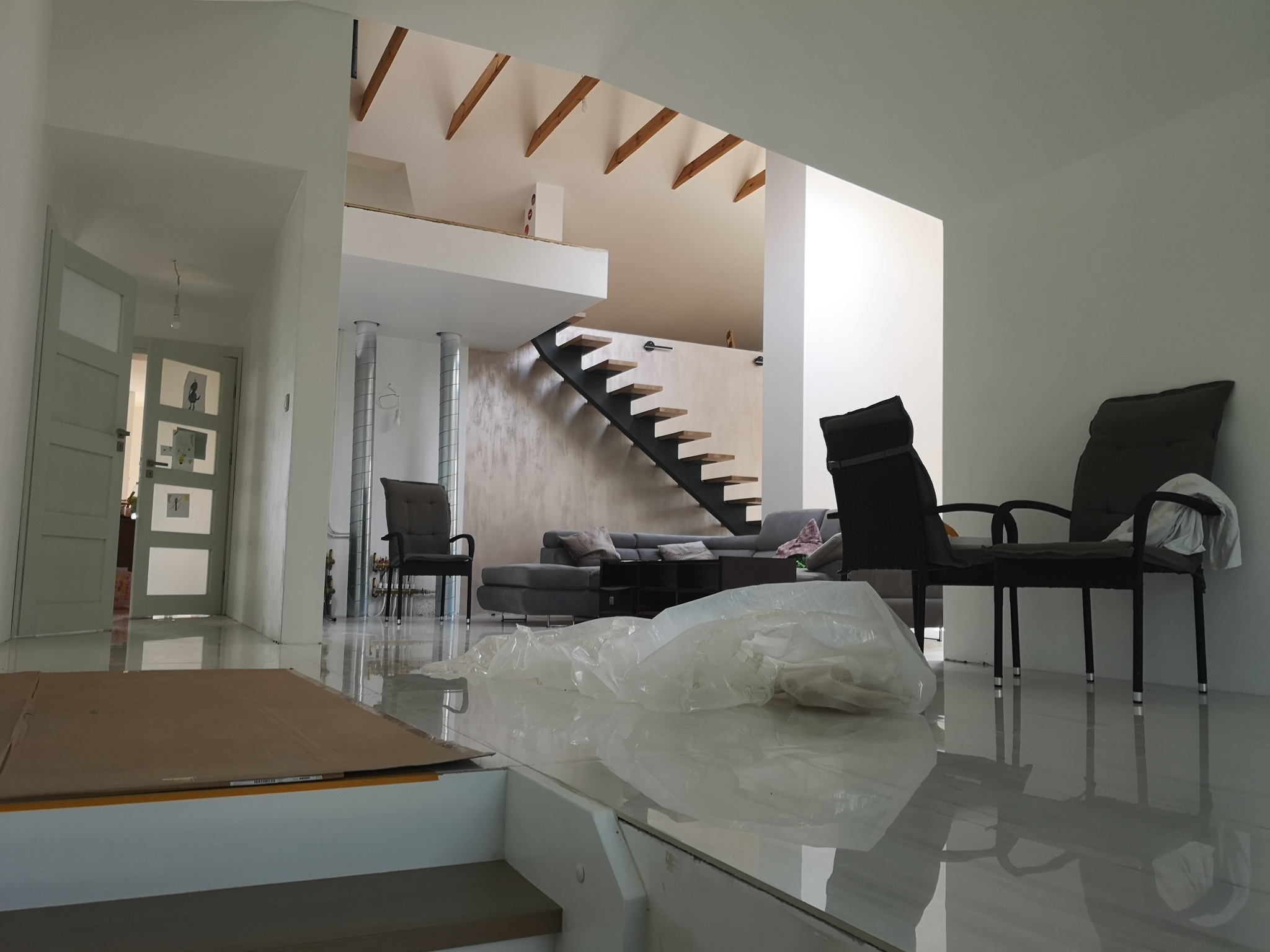 Wnętrze domu po remoncie: białe ściany, lśniąca podłoga z widocznymi refleksami, nowoczesne schody z drewnianymi stopniami i metalową konstrukcją, częściowo zasłonięte folią ochronną i kartonem,...