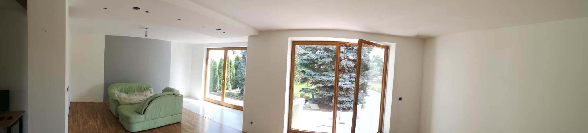 Panoramiczne ujęcie przestronnego, jasnego wnętrza w trakcie odświeżania, z widocznymi śladami prac malarskich na ścianach i suficie oraz dużymi oknami balkonowymi z drewnianą ramą, przez które...