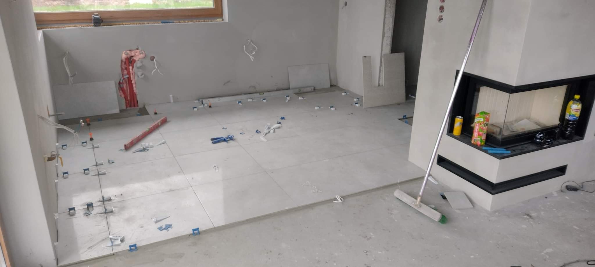 Układanie jasnych, wielkoformatowych płytek podłogowych w pomieszczeniu z kominkiem, widoczne narzędzia glazurnicze i system poziomowania płytek.