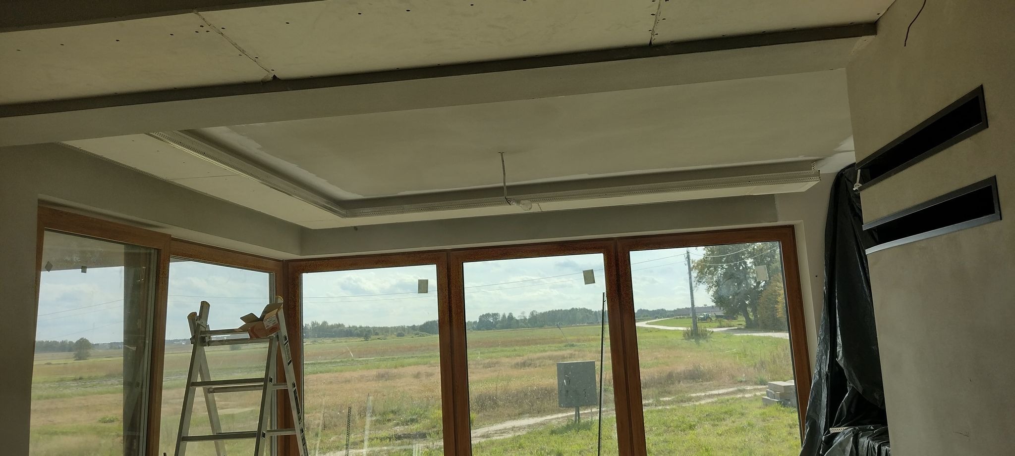 Wnętrze w trakcie wykańczania: sufit podwieszany z dekoracyjnym obramowaniem, widoczne otwory wentylacyjne w ścianie, okna z widokiem na pole, drabina oparta o okno.