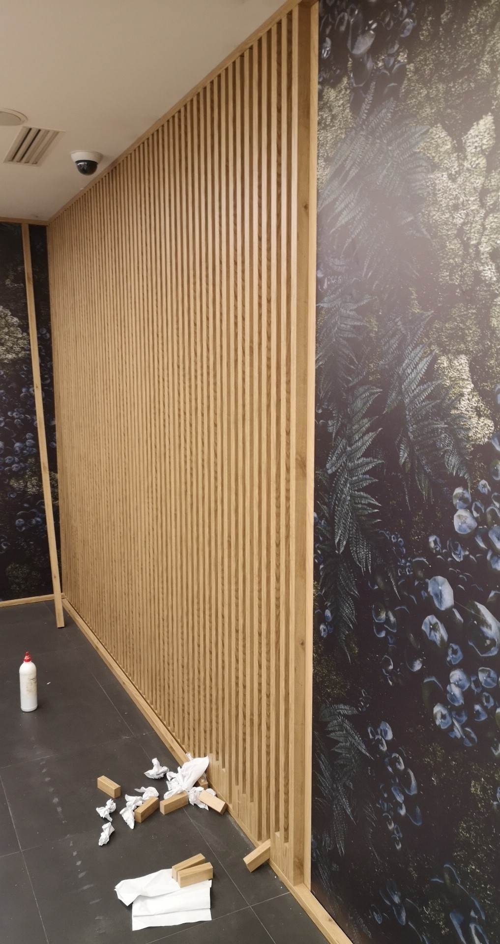 Montaż drewnianej ażurowej ścianki działowej na tle ściany z fototapetą o motywie leśnym, widoczne klocki dystansowe i resztki materiałów montażowych na podłodze.