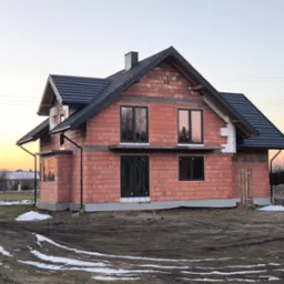 Surowy, dwukondygnacyjny budynek z czerwonej cegły z zamontowanymi oknami i ciemnym dachem w trakcie budowy, otoczony błotnistym terenem z resztkami śniegu i zachodzącym słońcem w tle.