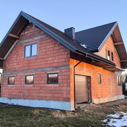 Z Natury Dobre - Surowy stan budynku z czerwonej cegły z zamontowanymi oknami i garażem, widoczny fundament i częściowo położony dach, z pozostałościami śniegu na trawie.