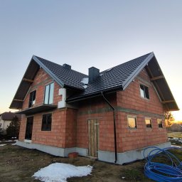 Z Natury Dobre - Surowy, dwukondygnacyjny dom z czerwonej cegły z czarnym dachem w trakcie budowy, widoczne okna bez szyb, fragmenty śniegu na ziemi i zwój niebieskiego węża ogrodowego.