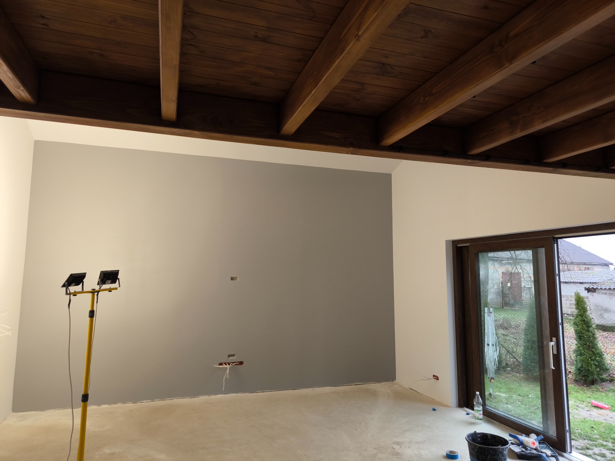 Wnętrze w trakcie prac wykończeniowych: pomalowana szara ściana, drewniany sufit z belkami, żółty statyw z lampami, widoczne kable elektryczne i okno przesuwne z widokiem na zewnątrz.