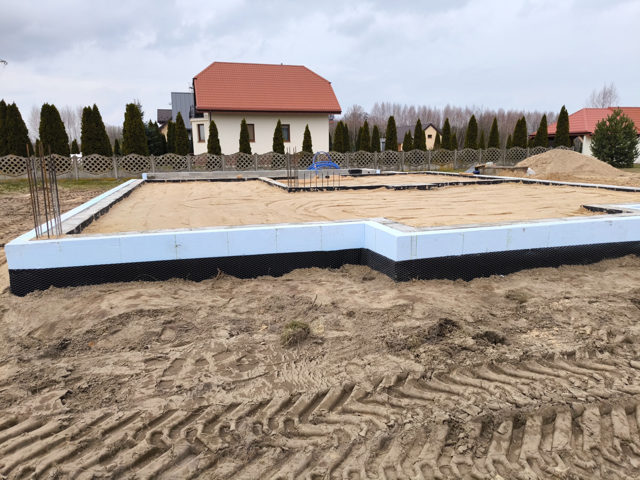 Przygotowany teren pod budowę domu z wylanym fundamentem, zabezpieczony izolacją termiczną i przeciwwilgociową, z widocznymi wystającymi prętami zbrojeniowymi.