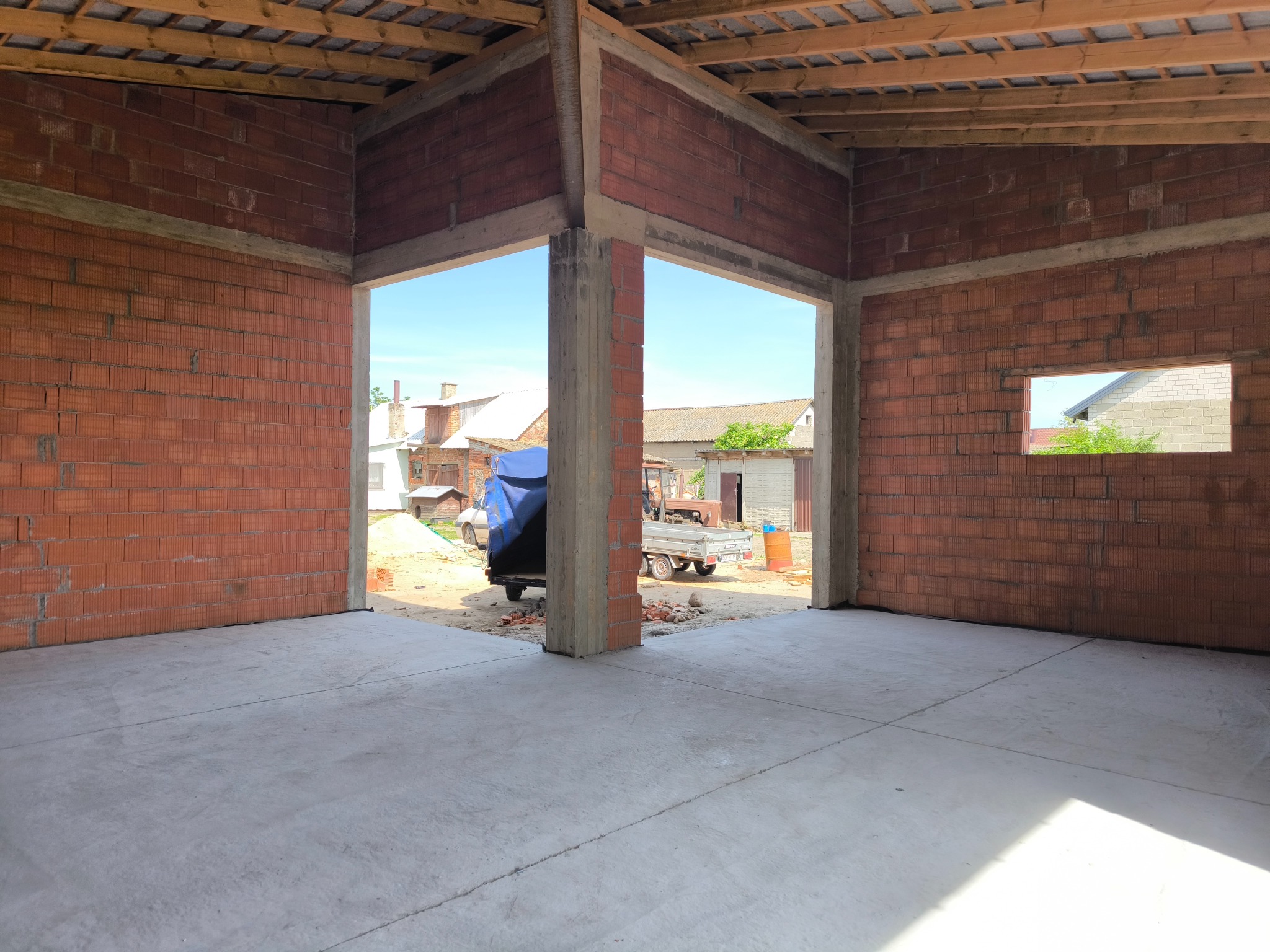 Wnętrze budynku gospodarczego w trakcie budowy: ściany z czerwonej cegły dziurawki, betonowa podłoga, widoczne elementy drewnianej konstrukcji dachu, widok na zewnątrz przez otwór drzwiowy...