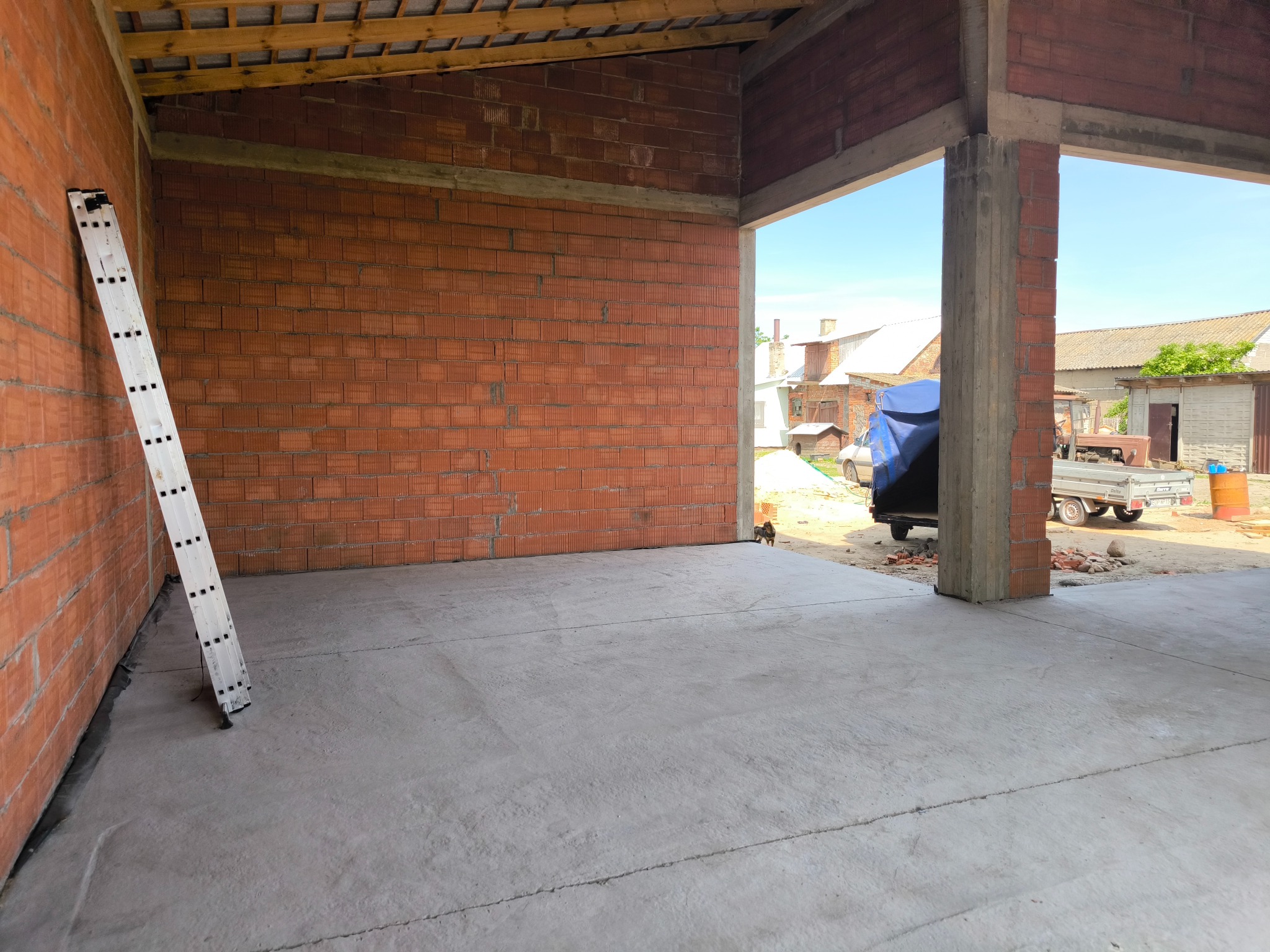 Wnętrze w surowym stanie budynku gospodarczego z czerwonej cegły, betonowa podłoga, drabina oparta o ścianę, widok na zewnątrz na posesję z przyczepą i budynkami gospodarczymi.