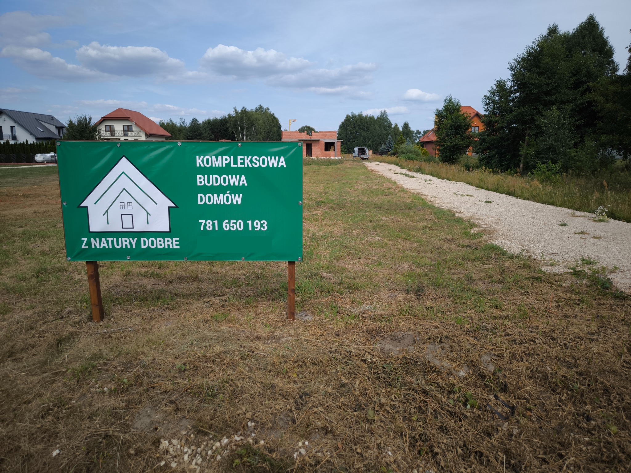Tablica reklamowa z informacją o kompleksowej budowie domów, w tle budowa domu w stanie surowym otwartym i droga gruntowa.
