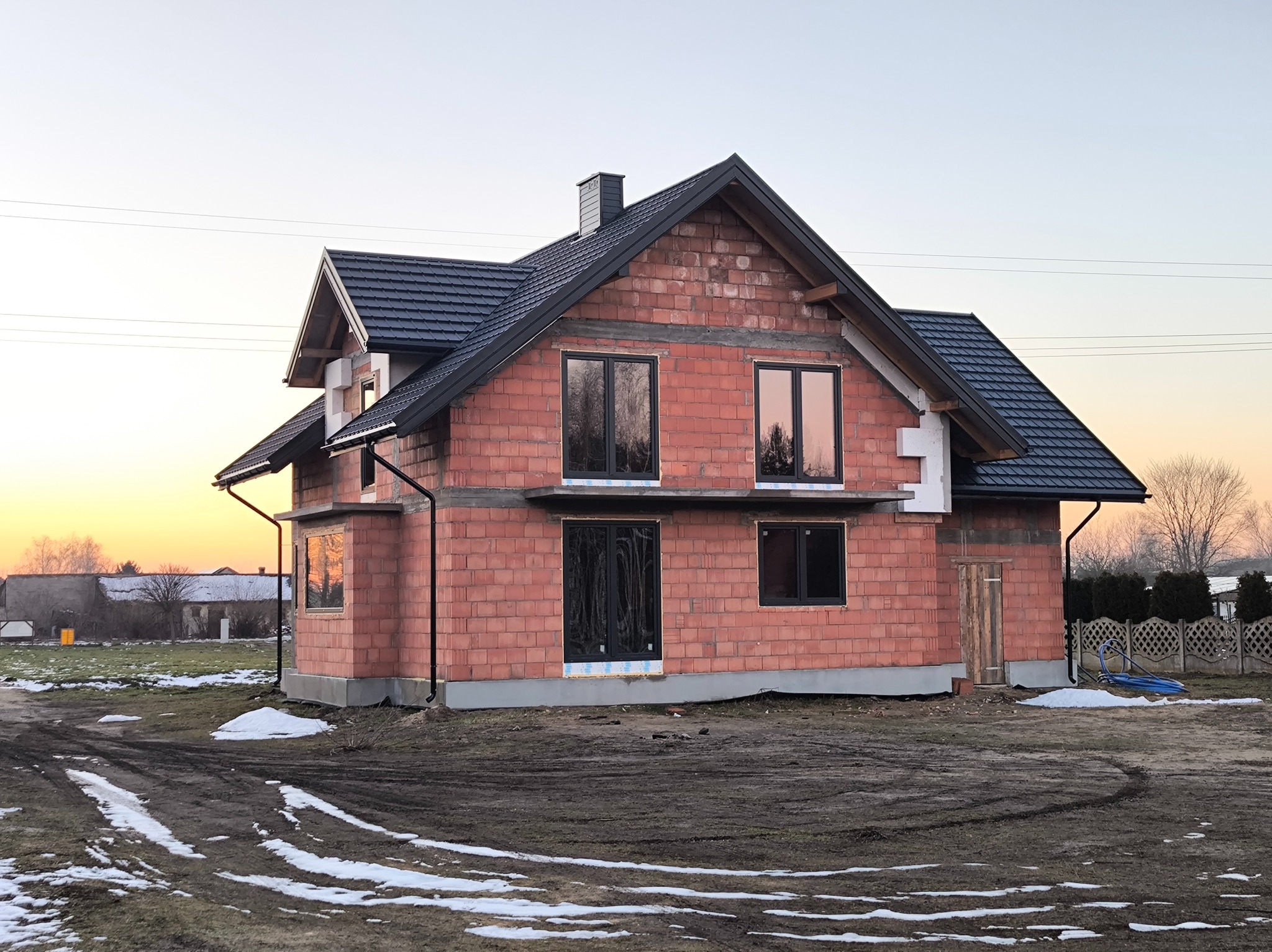 Surowy, dwukondygnacyjny budynek z czerwonej cegły z zamontowanymi oknami i ciemnym dachem w trakcie budowy, otoczony błotnistym terenem z resztkami śniegu i zachodzącym słońcem w tle.