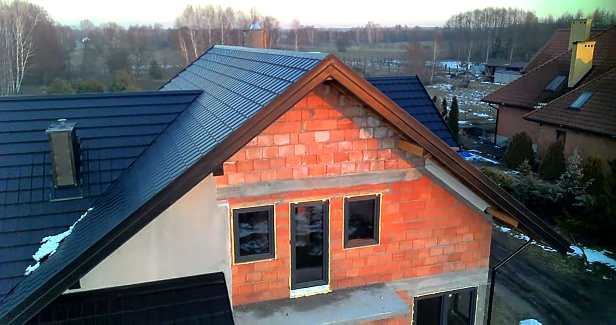 Wznoszenie domu z czerwonej cegły, widoczne wstawione okna, dach z ciemnej blachy, zimowy krajobraz w tle.