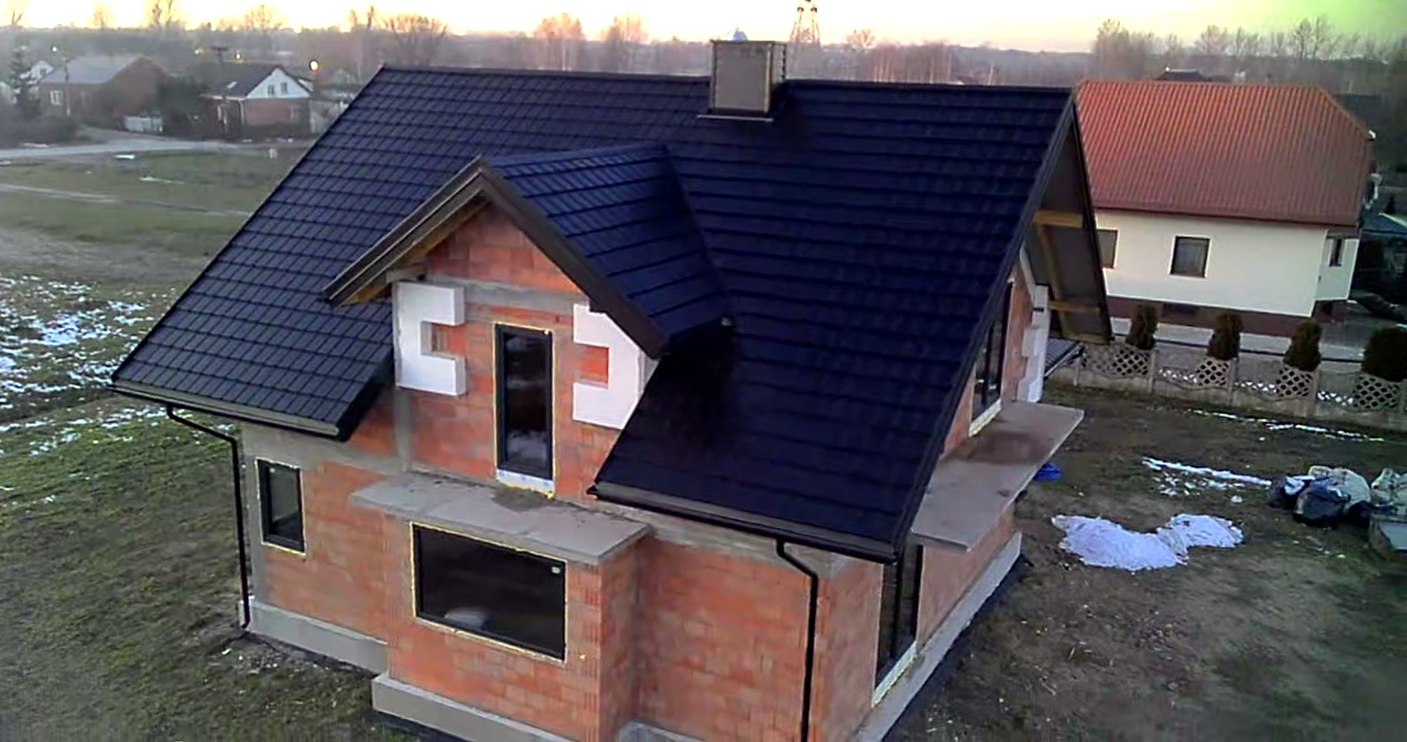 Niedokończony dom jednorodzinny z ciemną dachówką, widoczne pustaki, okna bez parapetów i fragmenty styropianu na elewacji, na zewnątrz rozłożone materiały budowlane.