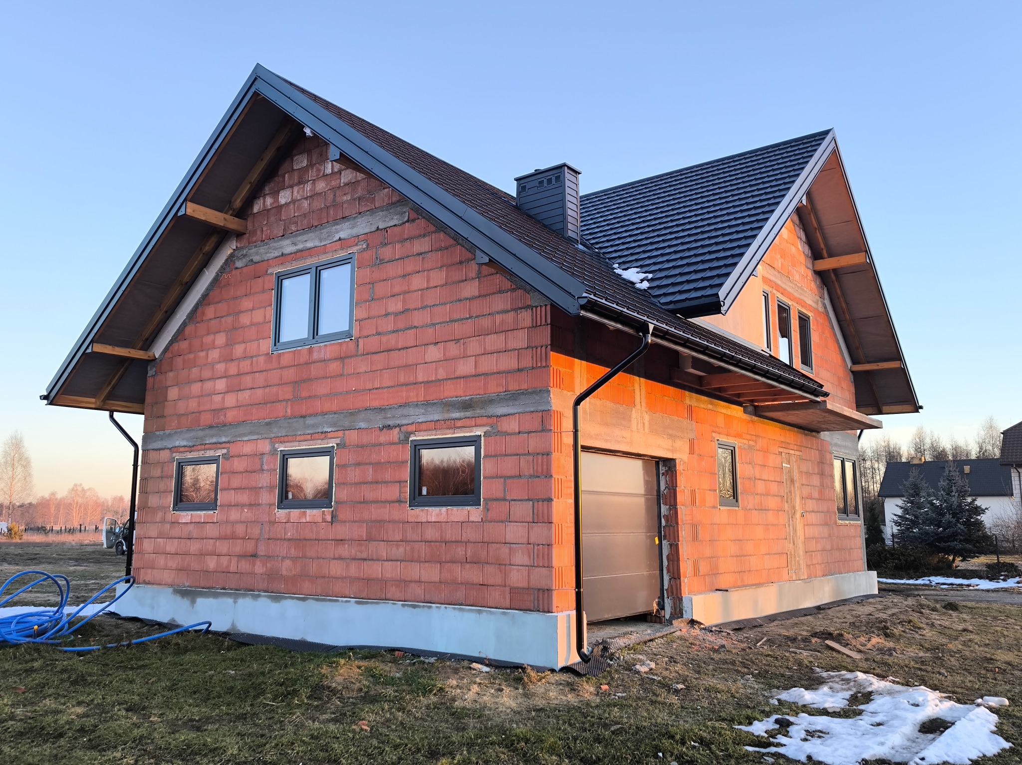 Surowy stan budynku z czerwonej cegły z zamontowanymi oknami i garażem, widoczny fundament i częściowo położony dach, z pozostałościami śniegu na trawie.