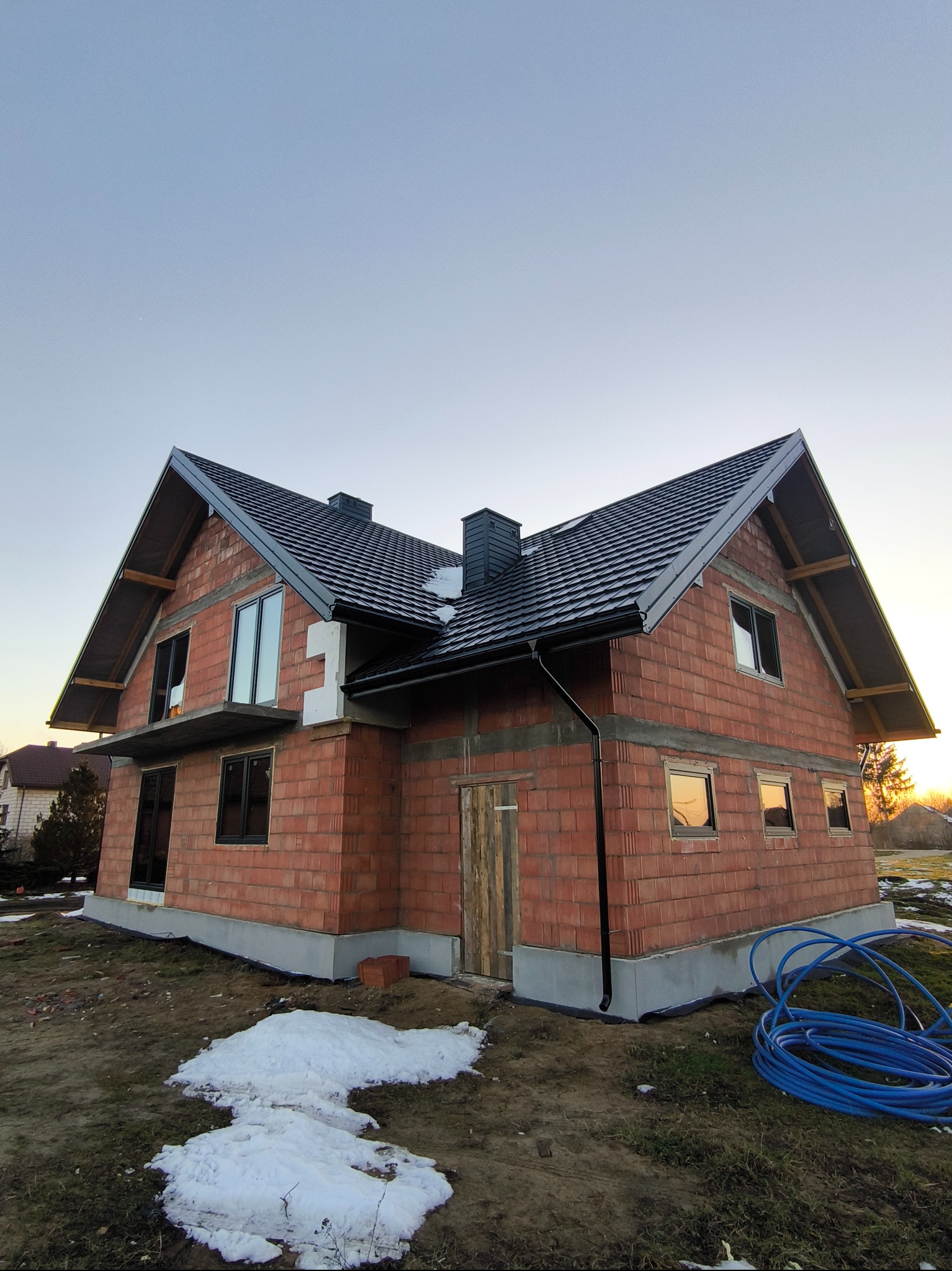 Surowy, dwukondygnacyjny dom z czerwonej cegły z czarnym dachem w trakcie budowy, widoczne okna bez szyb, fragmenty śniegu na ziemi i zwój niebieskiego węża ogrodowego.