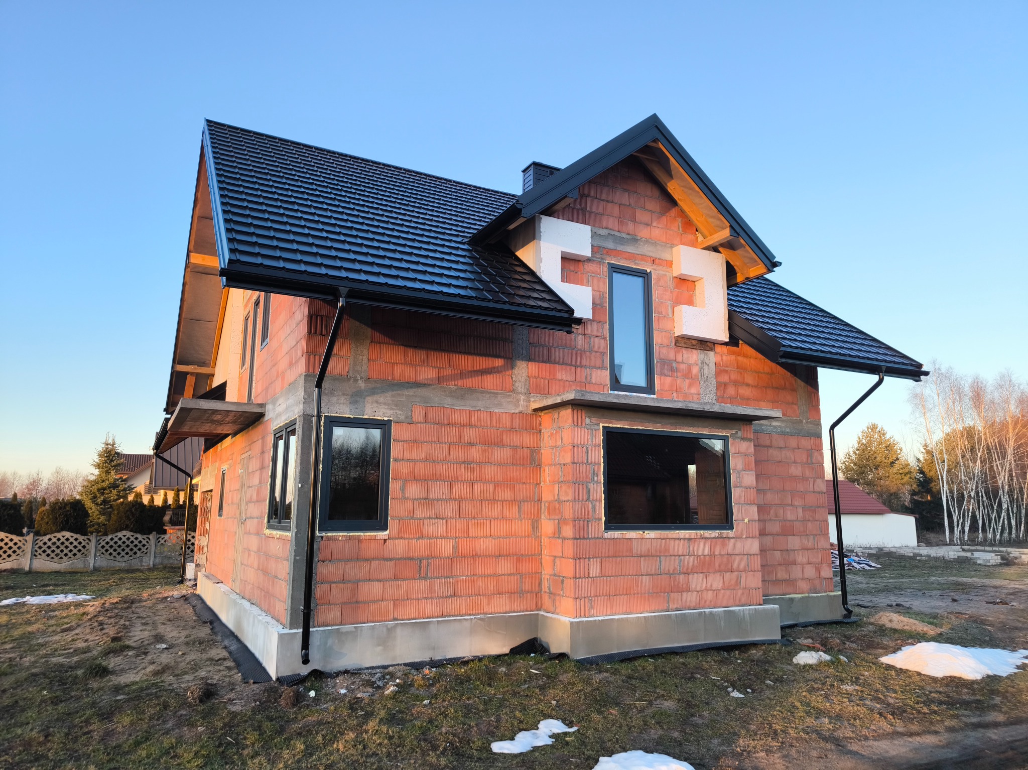 Surowy stan domu z czerwonej cegły z czarną dachówką i oknami, widoczne elementy izolacji wokół okien, krajobraz w tle.