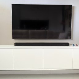 ESEN STUDIO - Biała szafka RTV z czterema drzwiczkami, soundbarem, szarą wazą i trzema miniaturowymi samochodzikami na blacie pod wiszącym telewizorem.