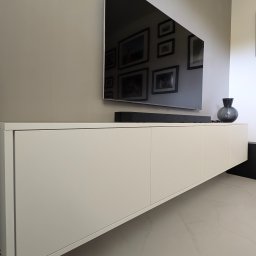 ESEN STUDIO - Minimalistyczna, biała szafka RTV wisząca pod telewizorem, z głośnikiem soundbar na blacie i szarą wazą, w salonie w Gdańsku. W odbiciu ekranu widoczne ramki ze zdjęciami na ścianie.