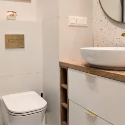 Nowoczesna łazienka z podwieszaną miską WC, umywalką nablatową na drewnianym blacie i lustrem w złotej ramie na tle ściany w stylu lastryko.