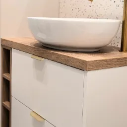 Nowoczesna szafka łazienkowa z umywalką nablatową i złotą armaturą, drewniany blat z p&oacute;łkami po lewej stronie, białe fronty szuflad z minimalistycznymi uchwytami, tło stanowi ściana z mozaiką.