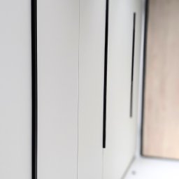 ESEN STUDIO - Białe, minimalistyczne drzwi szafy wnękowej z długimi, pionowymi, czarnymi uchwytami, widoczne z perspektywy bocznej, z rozmytym tłem jasnego pomieszczenia.