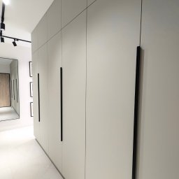 ESEN STUDIO - Długa, jasna szafa wnękowa z czarnymi, pionowymi uchwytami, widoczna w korytarzu z białymi marmurowymi płytkami i oświetleniem punktowym na suficie.