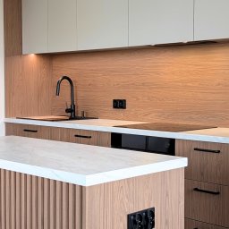 ESEN STUDIO - Nowoczesna kuchnia z drewnianymi frontami, białym blatem i czarną armaturą. Wyspa kuchenna z frezowanymi panelami. Oświetlenie LED pod szafkami.