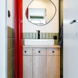 Nowoczesna łazienka z szafką pod umywalkę z drewnianymi frontami, białą umywalką nablatową, czarną armaturą i asymetrycznym lustrem. Czerwony grzejnik żeberkowy dodaje koloru. Widoczne zielone...