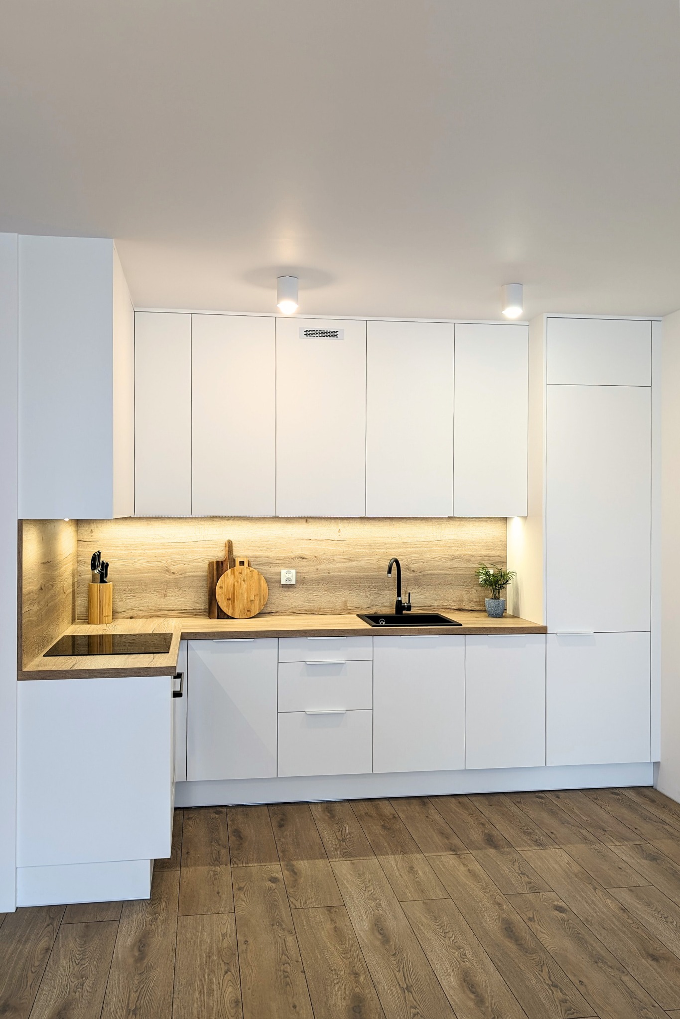 Nowoczesna biała kuchnia z drewnianym blatem i oświetleniem LED podszafkowym, czarna bateria kuchenna i drewniana podłoga.