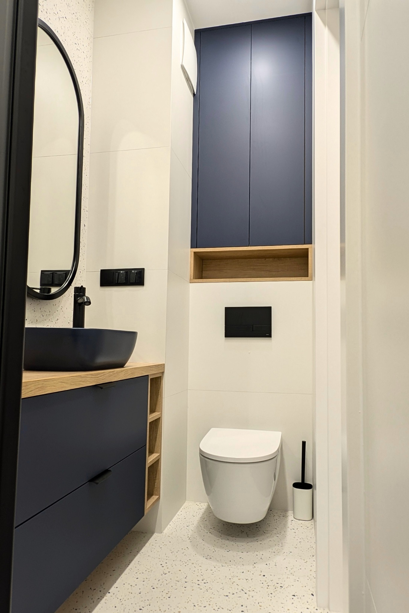Nowoczesna łazienka z zabudową meblową w odcieniach granatu i drewna, z umywalką nablatową i wiszącą toaletą, na podłodze lastryko.