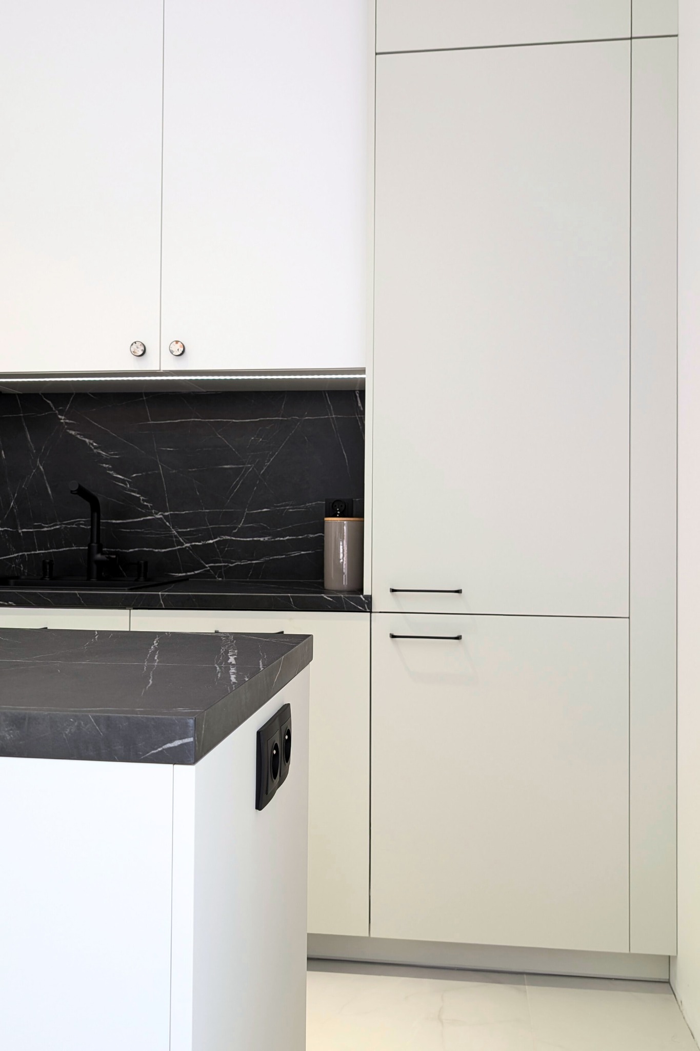 Minimalistyczna kuchnia: białe szafki z czarnymi uchwytami, blat i backsplash z czarnego marmuru, wyspa z gniazdkami elektrycznymi.