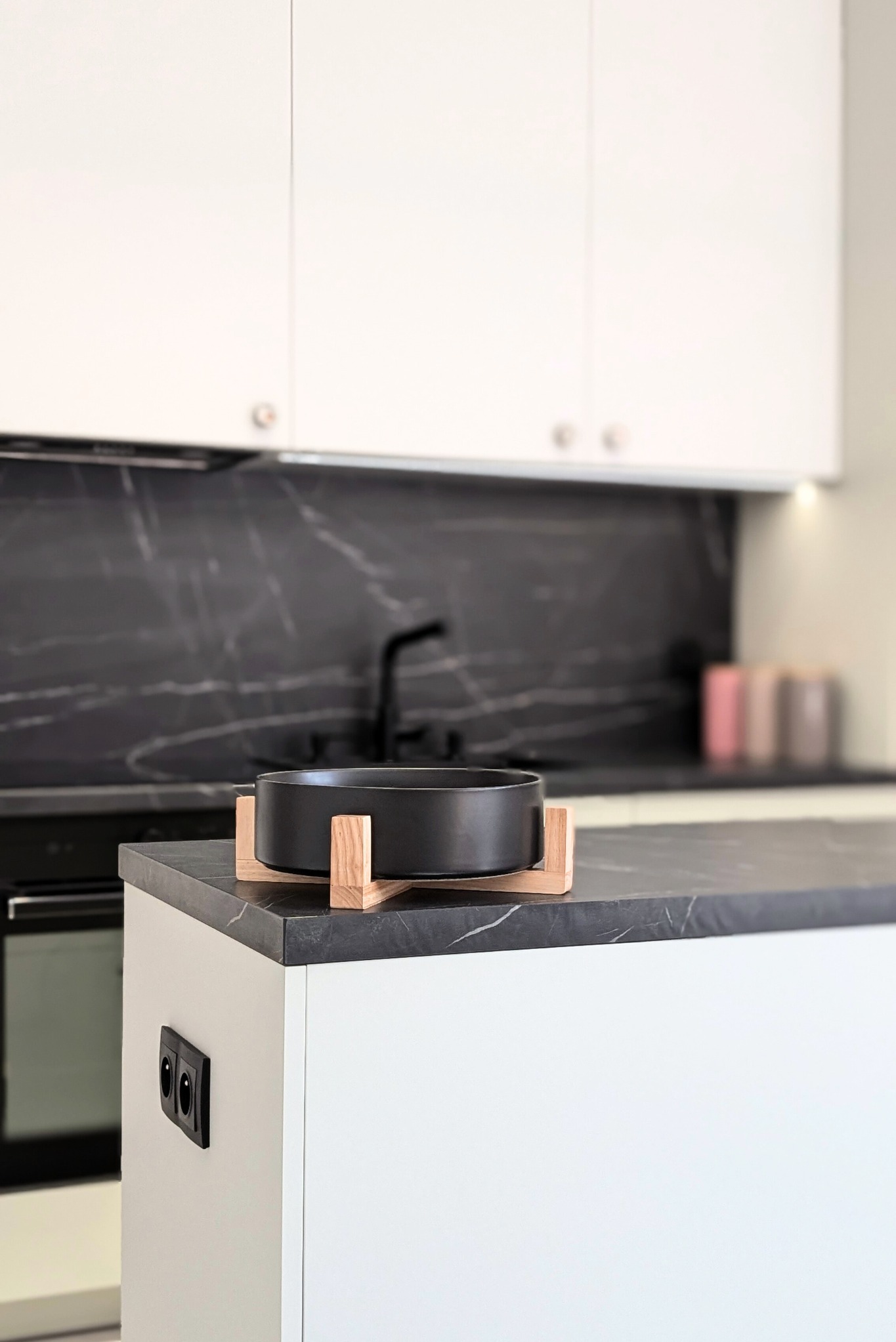 Nowoczesna kuchnia z białymi szafkami, czarnym blatem imitującym marmur i czarną misą na drewnianym stojaku na wyspie kuchennej. W tle widoczny czarny kran i szklane pojemniki.