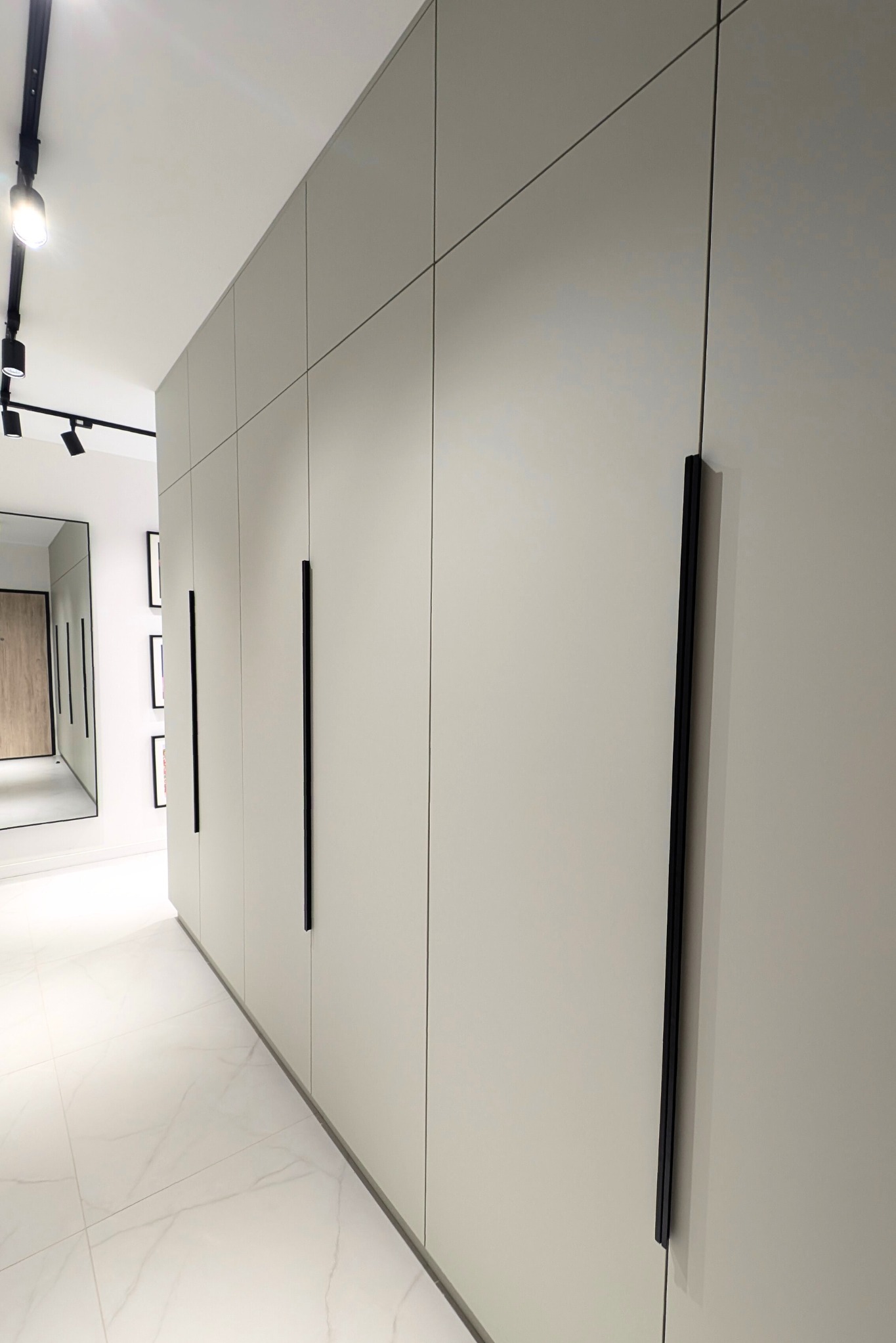 Długa, jasna szafa wnękowa z czarnymi, pionowymi uchwytami, widoczna w korytarzu z białymi marmurowymi płytkami i oświetleniem punktowym na suficie.