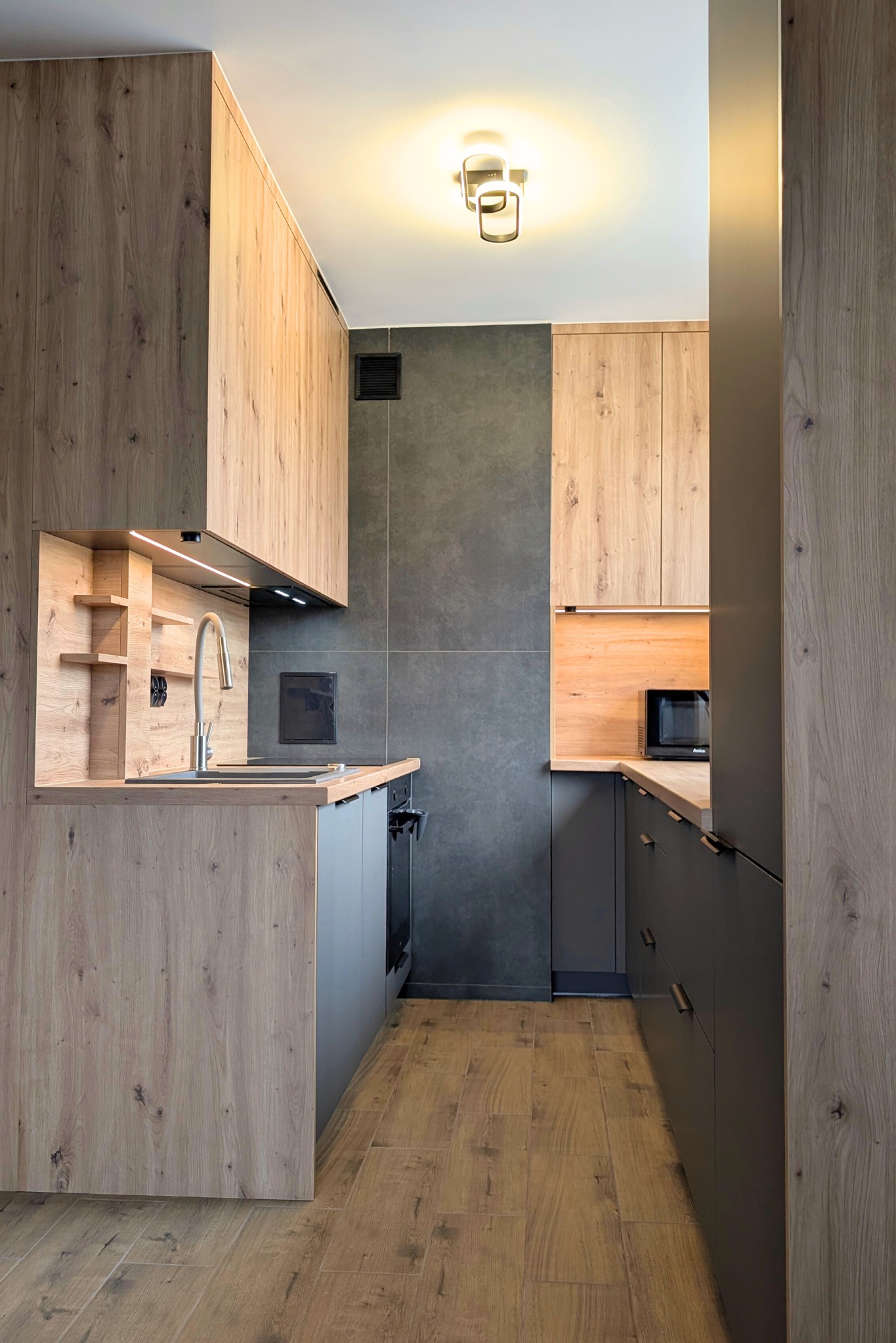 Wąska kuchnia w bloku z drewnianymi frontami szafek górnych i szarymi dolnymi. Ustawna zabudowa z oświetleniem LED i płytkami imitującymi beton.
