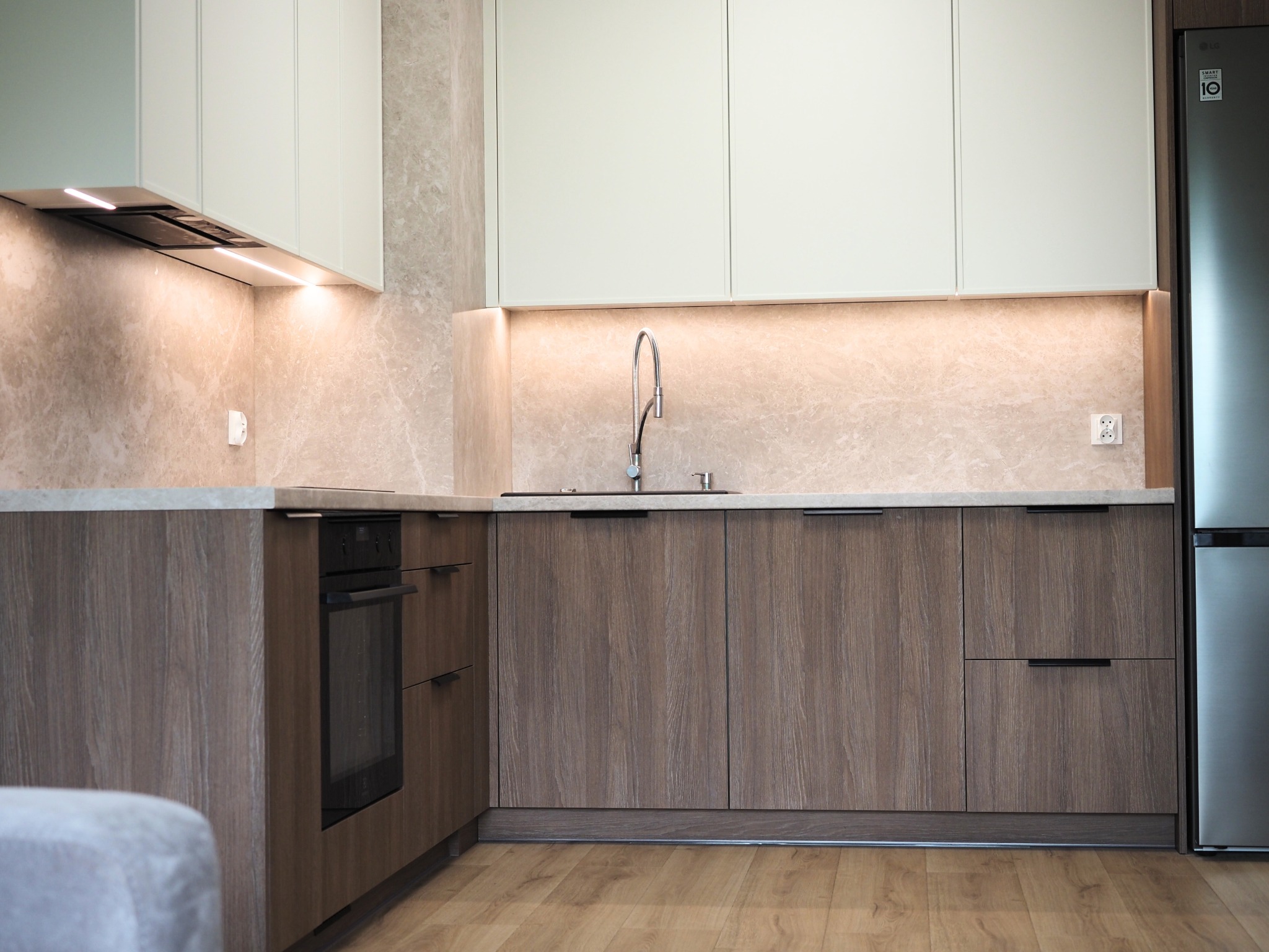 Jasna kuchnia z drewnianymi szafkami, oświetleniem LED pod szafkami wiszącymi i marmurowym blatem. Nowoczesny design z piekarnikiem i lodówką.