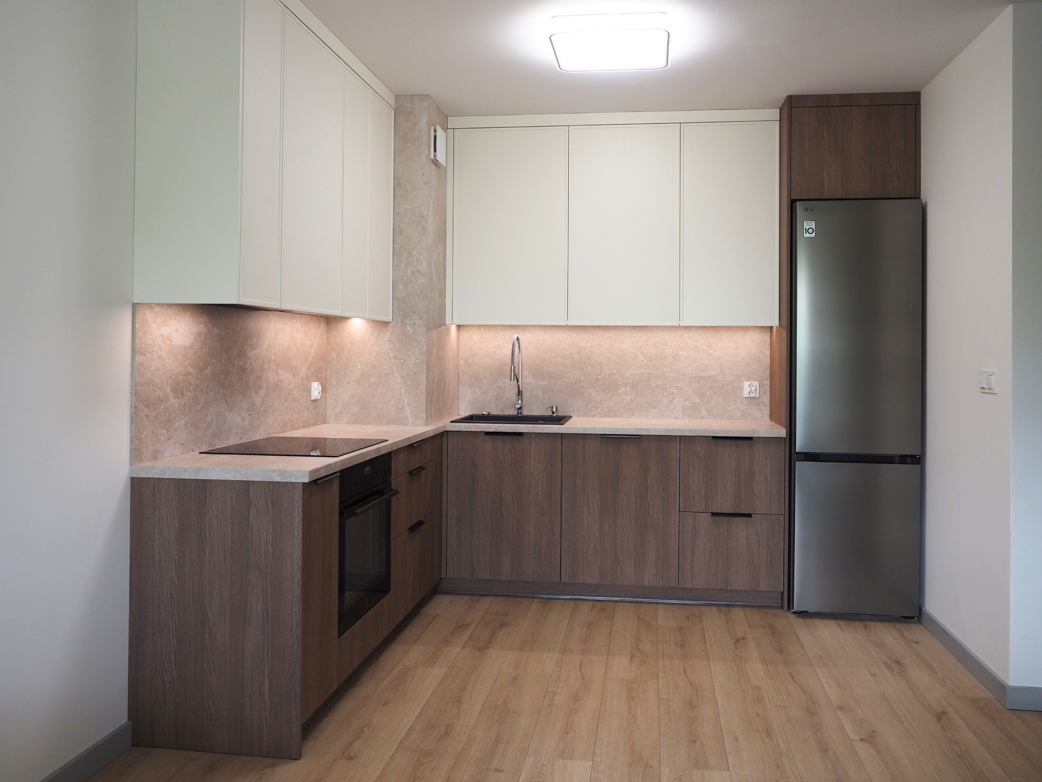 Nowoczesna kuchnia w kształcie litery L: jasne górne szafki, dolne z forniru drewna, marmurowy backsplash, czarny zlew i płyta, srebrna lodówka LG, parkiet.