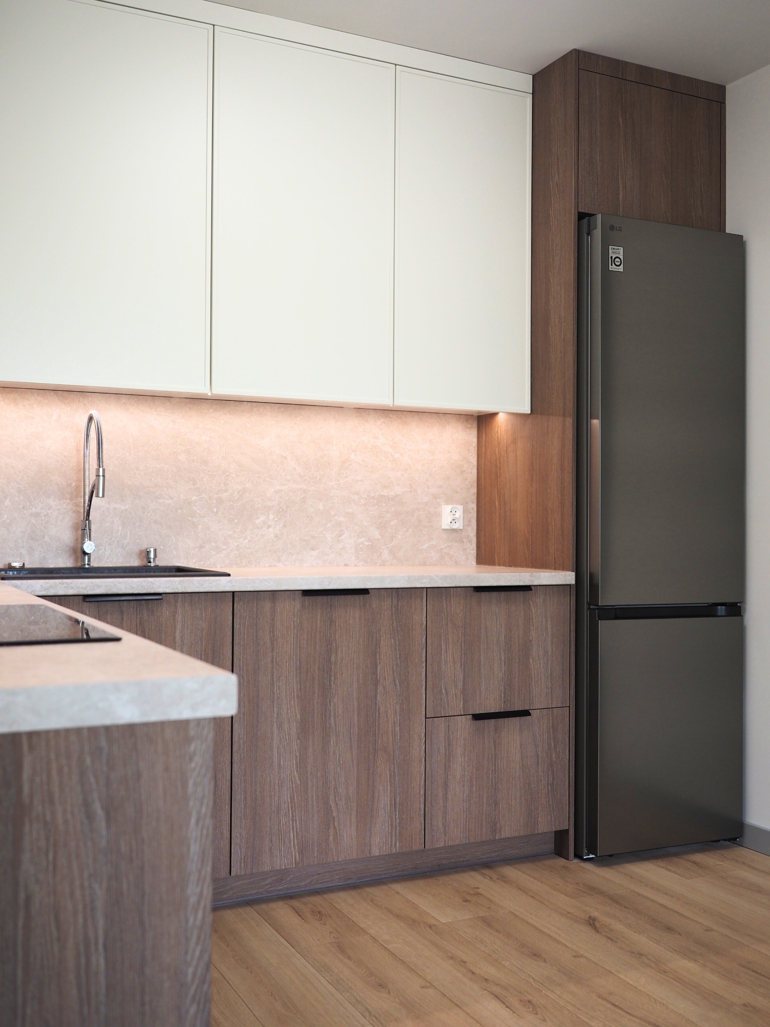 Nowoczesna kuchnia z drewnianymi szafkami dolnymi i białymi górnymi, oświetleniem LED pod szafkami i szarą lodówką LG. Minimalistyczny design.