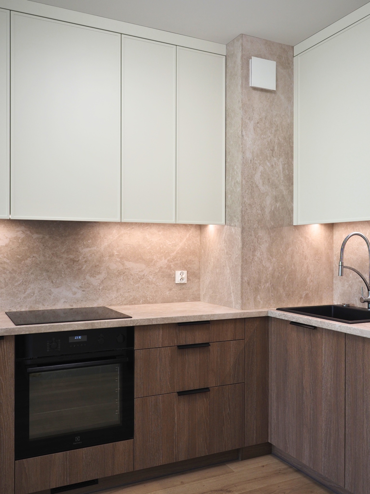 Kremowo-brązowa kuchnia narożna z oświetleniem LED pod szafkami, z czarnym zlewem i płytą indukcyjną. Fronty szafek z delikatnym wzorem drewna.