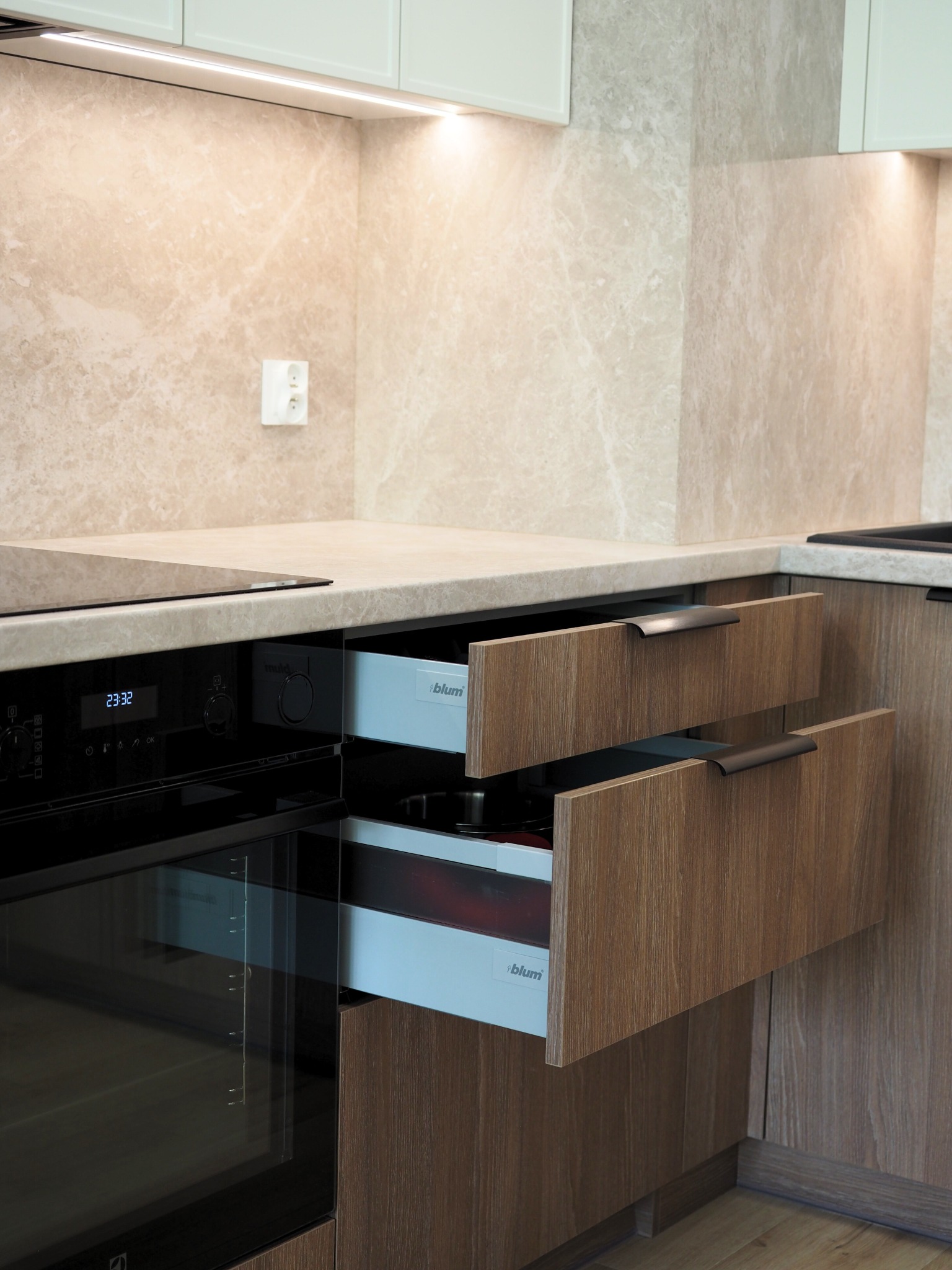 Nowoczesna zabudowa kuchenna z drewnopodobnymi frontami, wysuwanymi szufladami Blum i piekarnikiem Electrolux. Beżowy backsplash imitujący marmur, subtelne oświetlenie LED.