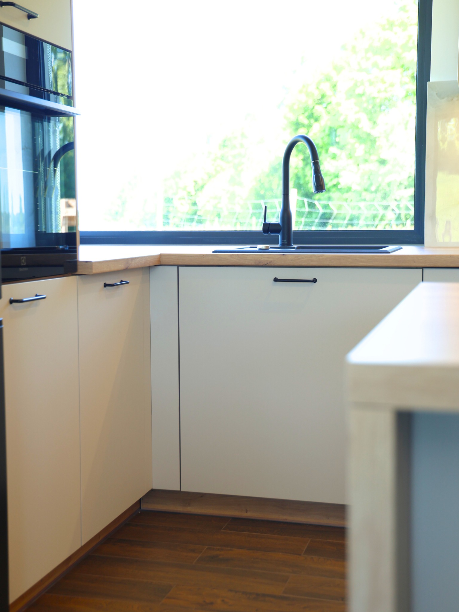 Jasne, minimalistyczne meble kuchenne z czarną armaturą i drewnianym blatem, na tle okna z widokiem na zieleń. Uwagę przykuwają długie, czarne uchwyty szafek.