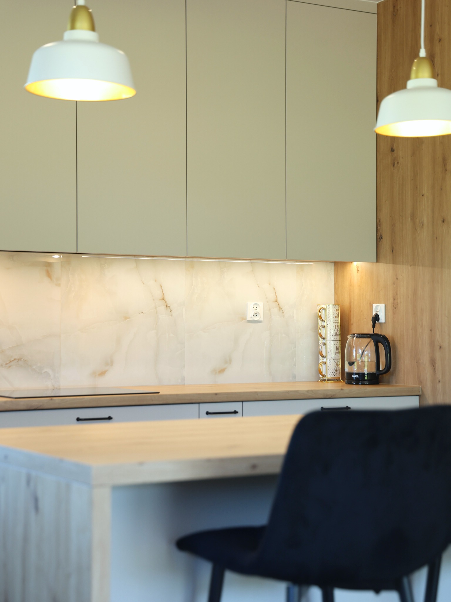 Nowoczesna kuchnia z oświetleniem LED pod szafkami, marmurowym panelem i drewnianym blatem. Widoczny czarny hoker i wiszące lampy.
