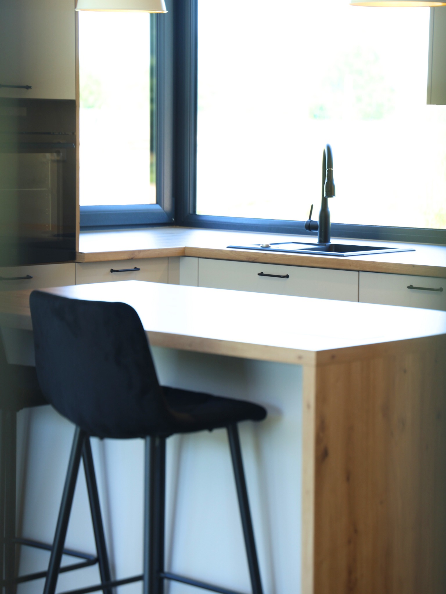 Jasna kuchnia z drewnianym blatem i czarną armaturą przy oknie. Krzesło barowe z czarnymi nogami. Nowoczesny design, minimalistyczne uchwyty.