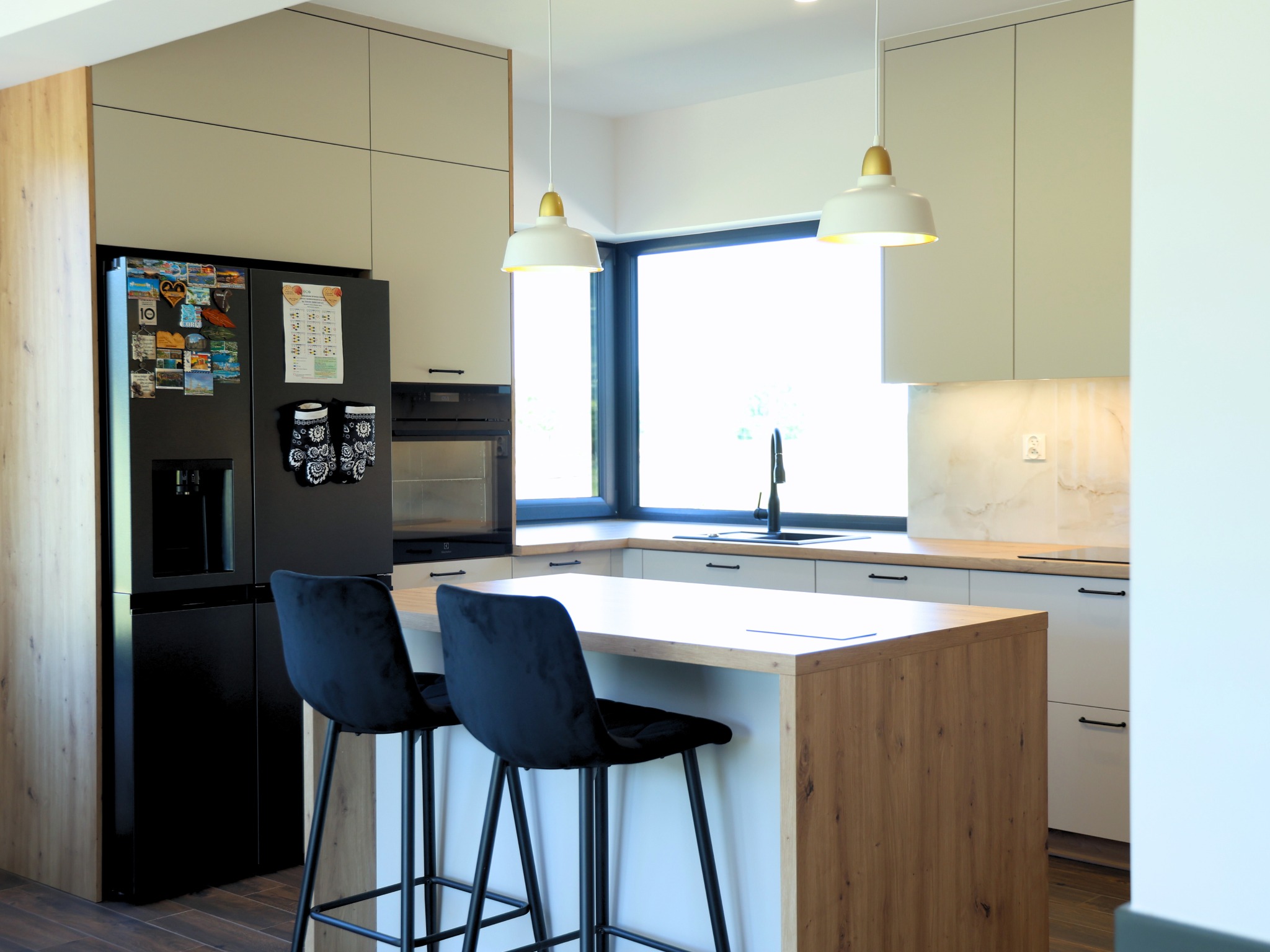 Nowoczesna kuchnia z wyspą i czarnymi hokerami. Jasne fronty szafek i drewniane akcenty. Na lodówce magnesy i organizer. Minimalistyczne lampy wiszące.