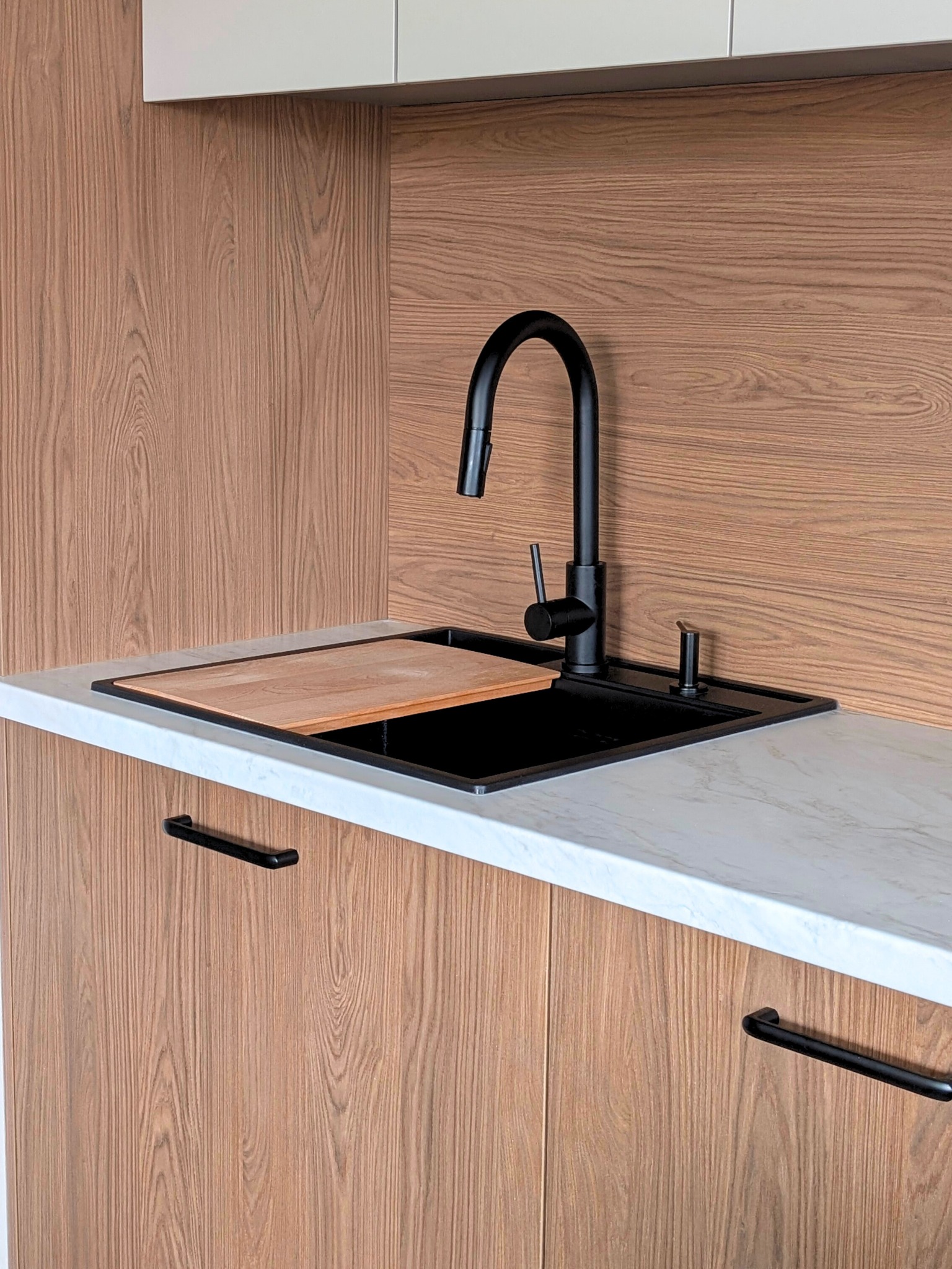 Nowoczesna kuchnia: czarny zlew z deską do krojenia, bateria i dozownik, drewniane fronty szafek z uchwytami, marmurowy blat. Minimalistyczny design.
