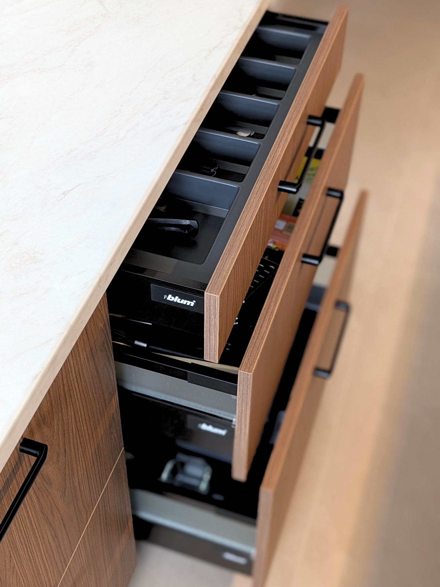 Otwarta, wysuwana szuflada kuchenna z przegródkami na sztućce i akcesoria, widoczny system Blum, drewniane fronty, blat marmurkowy.