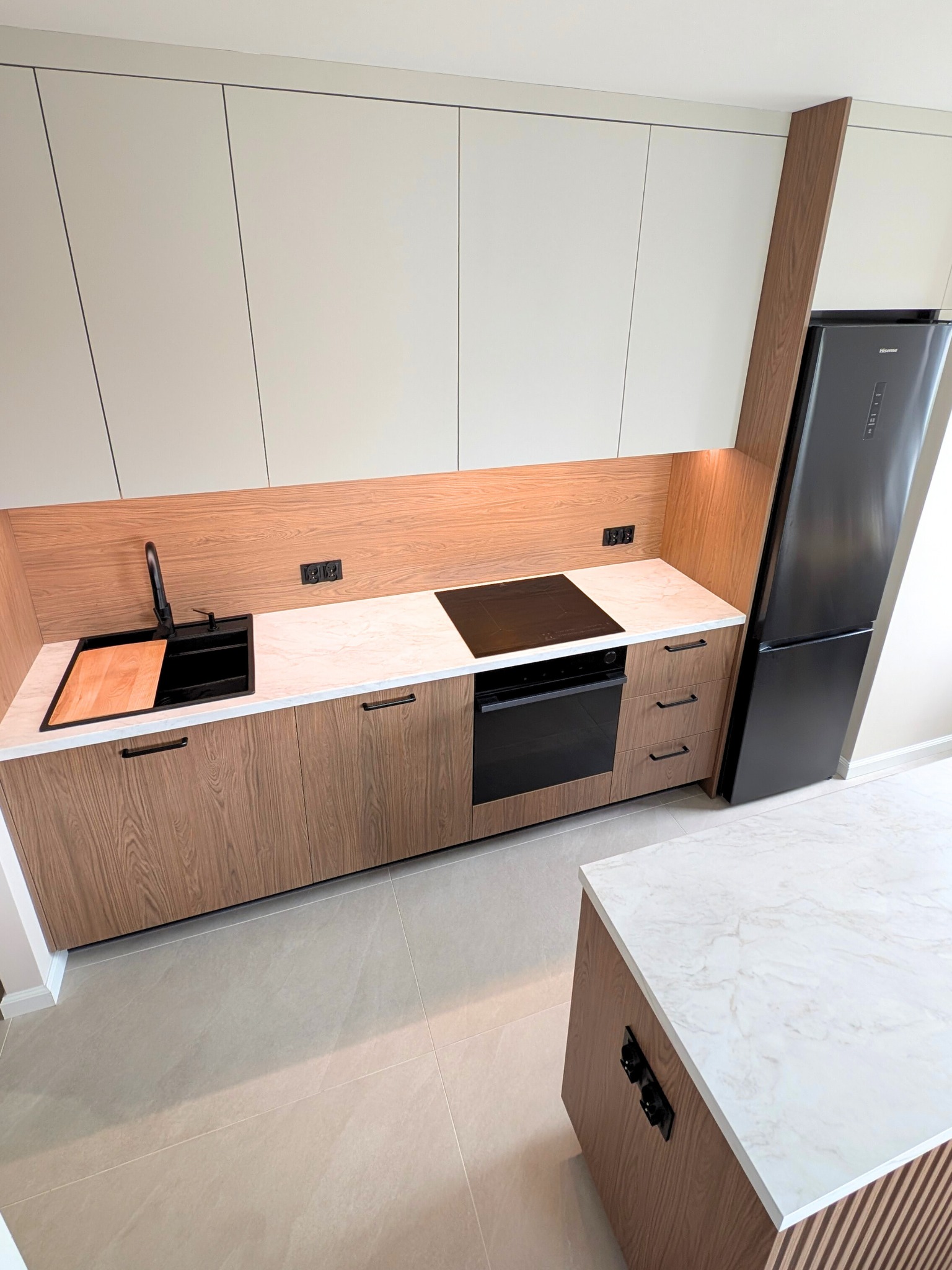 Nowoczesna kuchnia z drewnianymi frontami i białymi szafkami, czarnym zlewem i lodówką. Blat marmurkowy, oświetlenie punktowe, minimalistyczny design.