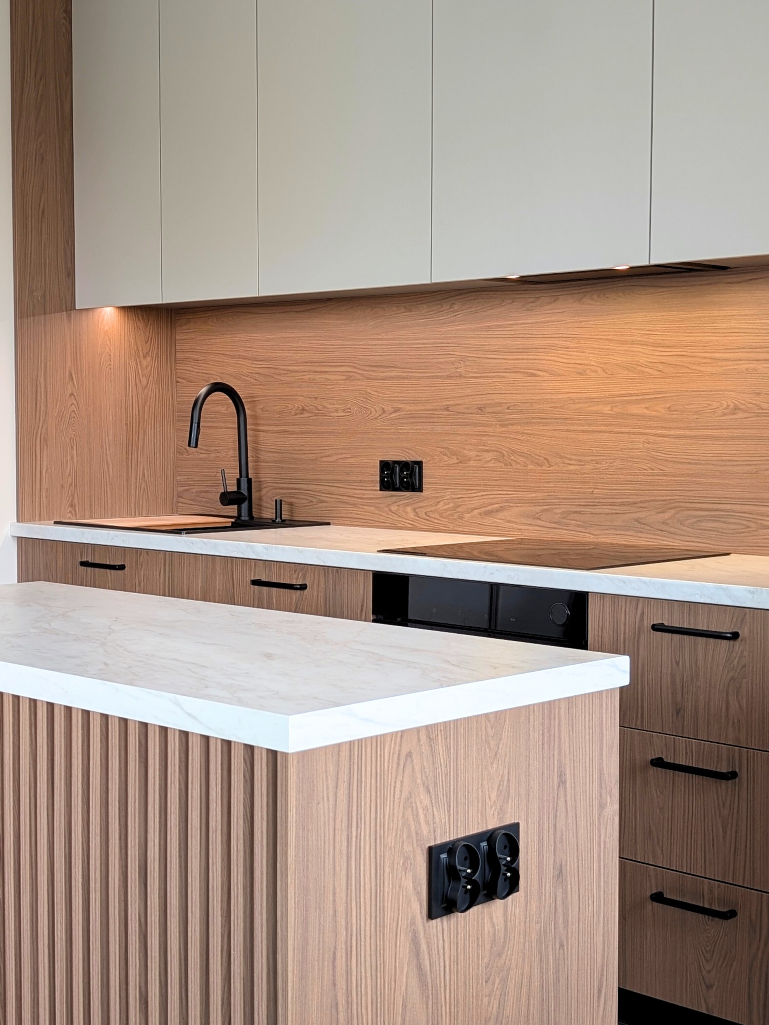 Nowoczesna kuchnia z drewnianymi frontami, białym blatem i czarną armaturą. Wyspa kuchenna z frezowanymi panelami. Oświetlenie LED pod szafkami.