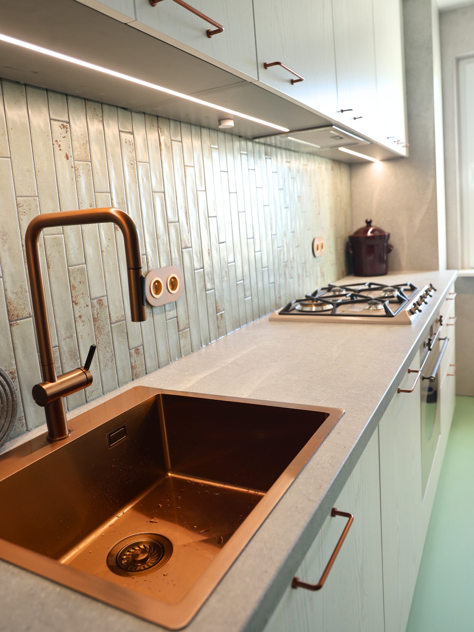 Nowoczesna kuchnia z miedzianym zlewem i baterią, jasne szafki z uchwytami w kolorze miedzi, płytki w stylu retro i oświetlenie LED pod szafkami.