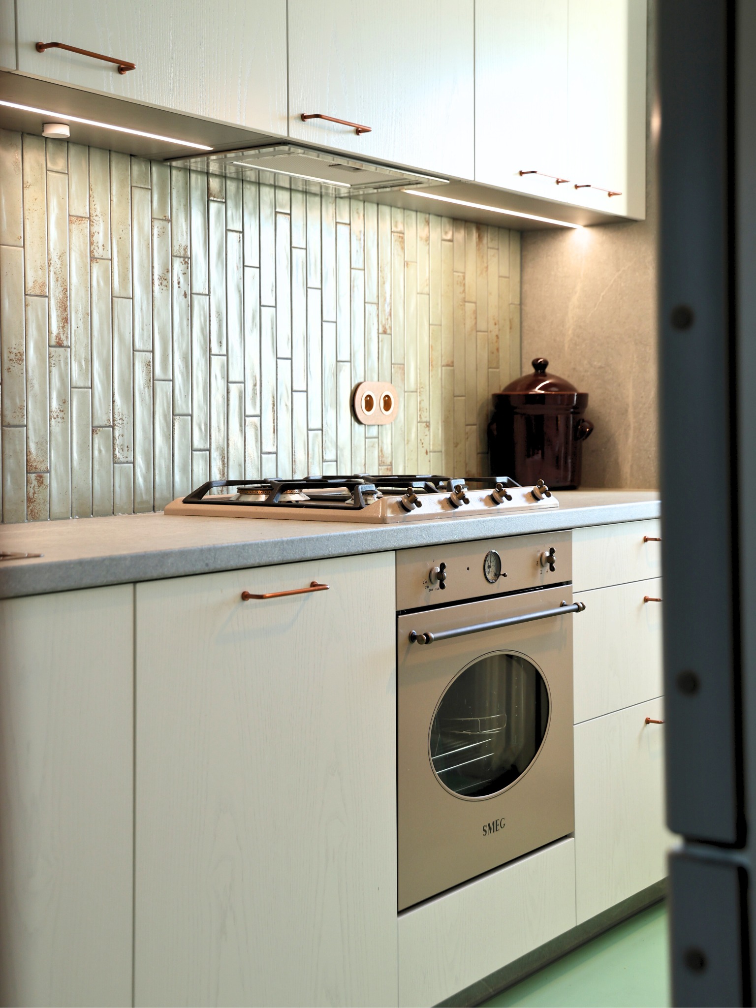 Jasna kuchnia z płytkami w stylu retro, miedziane uchwyty i piekarnik SMEG. Uwagę przykuwa ceramiczny garnek na blacie i nietypowe gniazdko.