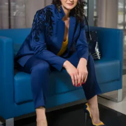 Kobieta w eleganckim, granatowym garniturze z aplikacjami na ramionach, siedząca na niebieskiej sofie i pozująca w żółto-granatowych szpilkach.