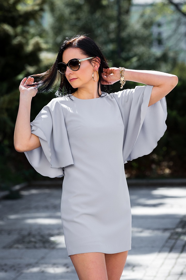 Kobieta w popielatej sukience z szerokimi rękawami, pozująca na zewnątrz w okularach przeciwsłonecznych i złotej bransoletce.